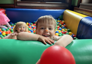 Na zdjęciu uśmiechnięta dziewczynka i chłopiec bawią się w basenie z kolorowymi, plastikowymi piłeczkami.