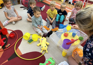 Na zdjęciu dzieci siedzą na dywanie i obserwują eksperyment pokazywany przez nauczycielkę.