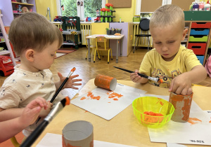 Na zdjęciu dwoje dzieci maluje na pomarańczowo rolki po papierze toaletowym.