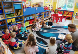 Na zdjęciu widać grupę dzieci siedzącą na dywanie w klasie oraz tatę jednego z dzieci, który czyta dzieciom książkę