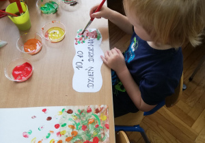 Chłopiec maluje kolorowe listki na kartce z napisem "Dzień Drzewa"