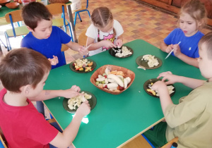 dzieci siedzą przy stoliku, kroją plastikowy nożykami różne owoce