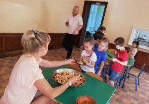 dzieci stoją w kolejce przed nauczycielka, która nakłada dzieciom sałatkę owocową przygotowana przez dzieci