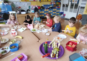 Dzieci siedzące przy stolikach wykonują owocowe szaszłyki i warzywne kukiełki.