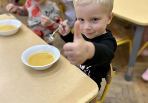 Na zdjęciu chłopiec podczas obiadu z zupą dyniową pokazujący podniesiony do góry kciuk.
