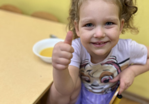 Na zdjęciu dziewczynka podczas obiadu z zupą dyniową pokazująca podniesiony do góry kciuk.