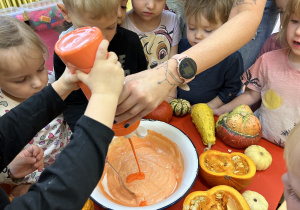 Na zdjęciu nauczycielka wraz z dziećmi miesza z misce pomarańczową masę do wykonania pracy plastycznej.