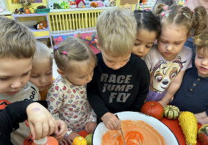 Na zdjęciu nauczycielka wraz z dziećmi miesza z misce pomarańczową masę do wykonania pracy plastycznej.