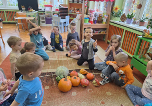 Na zdjęciu siedzą dzieci na dywanie, porównują wielkość, kolor i rodzaje dyń.