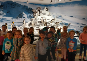 na zdjęciu grupa dzieci przy interaktywnym dużym globusem, w tle ekspozycja antarktycznego środowiska i eksponaty - pingwiny