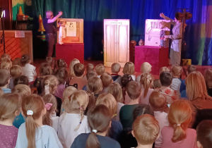 na zdjęciu dzieci oglądają przedstawienie w tle dwoje aktorów trzymających lalki - marionetki chłopca i psa