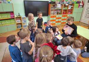 Mama Julci czyta bajkę dzieciom siedzącym na dywanie i pokazuje ilustracje w niej zawarte.