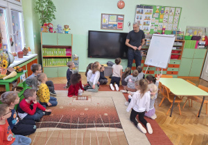 Na zdjęciu tata Iskry – wykonujący zawód architekta- dzieciom siedzącym na dywanie, pokazuje na tablicy narysowany dom.