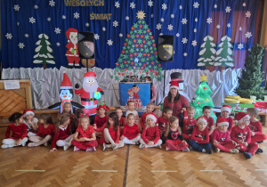 Na zdjęciu dzieci z grupy Skrzaty z wychowawcą, w tle na kotarze znajduje się dekoracja zimowa na której jest Mikołaj i choinka.