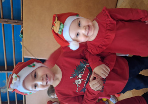 Na zdjęciu chłopiec i dziewczynka ubrani na czerwoni w czapkach Mikołaja, uśmiechają się do zdjęcia.