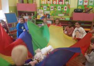 Zdjęcie przedstawia dzieci podczas zabawy ruchowej przy muzyce z wykorzystaniem chusty animacyjnej i pluszaków