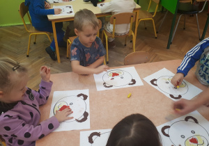 Zdjęcie przedstawia dzieci siedzące przy stolikach w trakcie realizacji pracy plastycznej "Mój miś” – etap pierwszy - wyklejanie elementów szablonu plasteliną