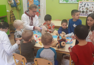 zdjęcie przedstawia dzieci i studentów siedzące przy stolikach podczas zabawy z wykorzystaniem pluszowych maskotek i zabawkowych akcesorii medycznych
