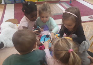 zdjęcie przedstawia dzieci i studentów siedzące przy stolikach podczas zabawy z wykorzystaniem pluszowych maskotek i zabawkowych akcesorii medycznych