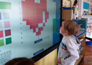 Dziecko stoi przed monitorem interaktywnym i patrzy na dyktando graficzne przedstawiające serce