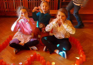 Na podłodze kontur serca ułożony z czerwonych kubeczków. W środku siedzą trzy dziewczynki pokazując serca ułożone z dłoni