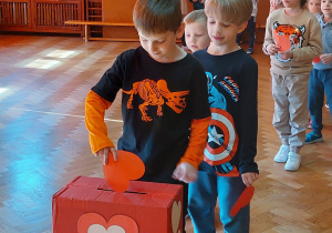 Dzieci stoją z kartkami walentynkowymi w rzędzie, po kolei wrzucają kartki do pudełka ozdobionego serduszkami - walentynkowej skrzynki