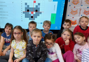 Dzieci siedzą przed tablicą interaktywną, na której widnieje rozwiązane dyktando graficzne przedstawiające kota.