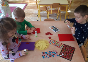 zdjęcie przedstawia dzieci z grupy ,,Misie’’ siedzące przy stolikach podczas wykonywania pracy plastycznej ,,Kartka walentynkowa” z wykorzystaniem dostępnych materiałów plastycznych